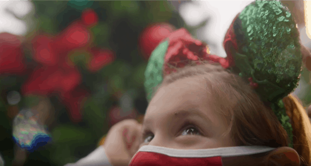 A young Disney fan in a festive red mask and sparkling green and red Mickey ears gazes up, surrounded by the shimmering lights of a Christmas tree at Walt Disney World, celebrating holiday magic in true park style.