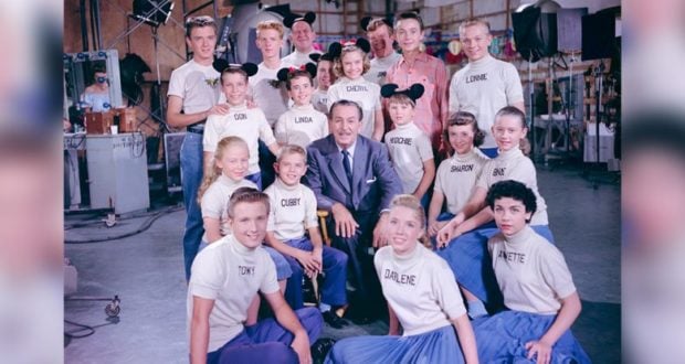 Fans of Disney entertainment gather in a vibrant TV studio, some sporting signature Mickey Mouse ears and custom shirts with their names, as they join in the festivities honoring 65 magical years of the beloved Mickey Mouse Club show.