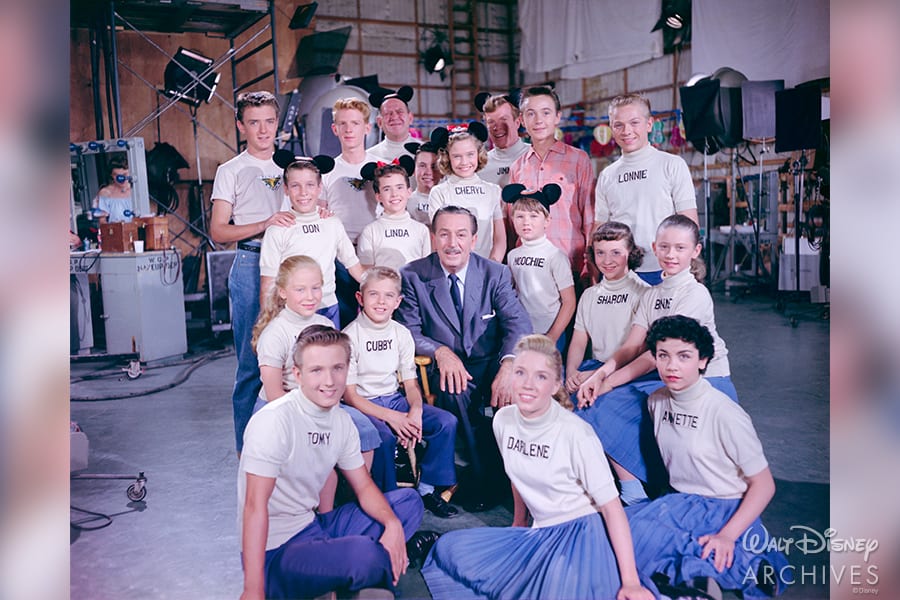 Walt Disney joins the original Mouseketeers, all sporting iconic name shirts and mouse ears, inside a studio set—a nostalgic tribute to 65 years of the beloved Mickey Mouse Club that shaped Disney entertainment history.