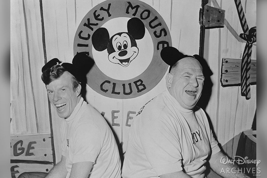 Disney fans sporting Mickey Mouse ears sit back-to-back, beaming in front of the iconic Mickey Mouse Club sign, commemorating 65 magical years of Disney entertainment.