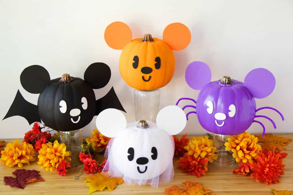Four Disney character pumpkins with signature mouse ears, inspired by favorite park icons and painted for Halloween, are displayed as a festive centerpiece amid autumn leaves and flowers, bringing magical Disney Parks decor to your celebration.