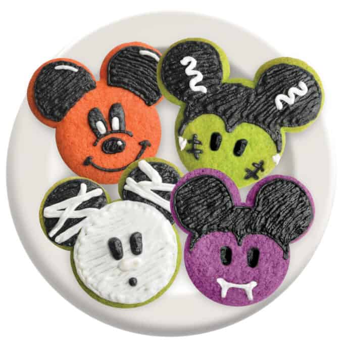 Four vibrant cookies shaped like Mickey and Minnie Mouse heads, each decorated to resemble iconic Disney Halloween characters, are artfully arranged on a white plate—an enchanting treat for any Disneyland or Walt Disney World Halloween celebration.