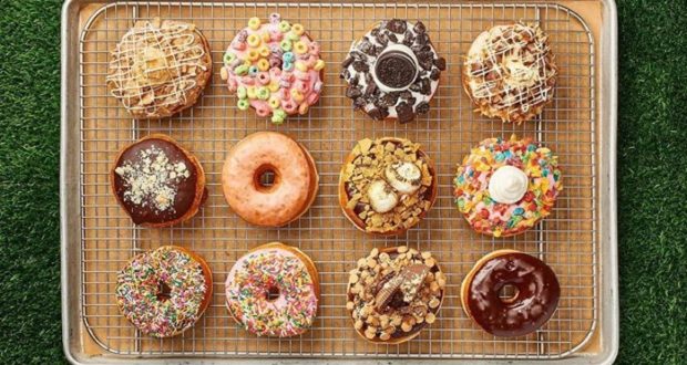 A colorful assortment of Disney Springs donuts, each topped with playful and imaginative decorations, sits on a wire rack atop a tray, ready for guests to enjoy on the lush green grass during a magical day at Walt Disney World Resort.