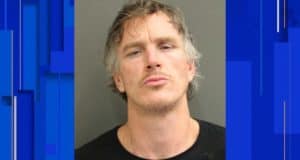 A park employee in character, sporting tousled hair and a dramatic expression, poses for a themed mugshot against a simple backdrop as part of an immersive attraction experience.