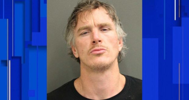 A park employee in character, sporting tousled hair and a dramatic expression, poses for a themed mugshot against a simple backdrop as part of an immersive attraction experience.