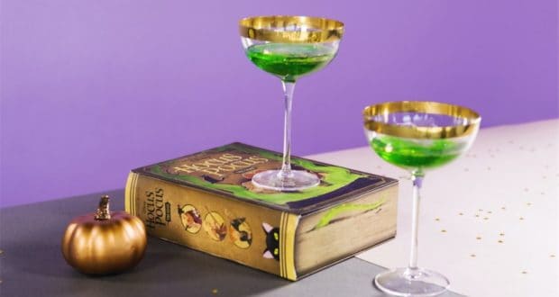 Two enchanted green drinks, crafted to resemble magical potions, are served beside a Hocus Pocus spell book and a shimmering gold pumpkin, all set against a vibrant purple backdrop straight out of a Halloween celebration at a Disney theme park.