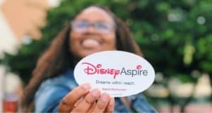 A Cast Member at Disney World beams while holding up a Disney Aspire sticker that says, “Dreams within reach. Aspire.Disney.com,” highlighting the chance for fellow Cast Members to further their education for free.