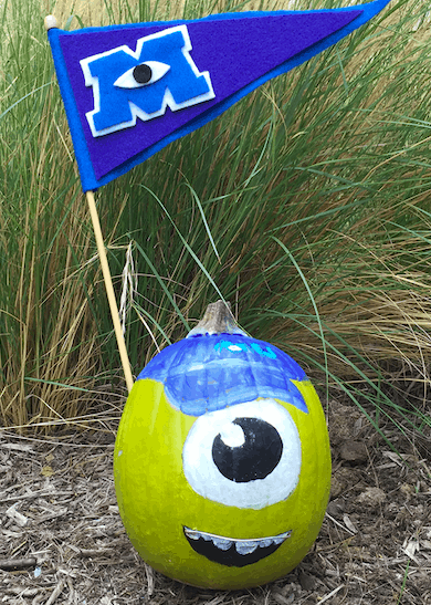 Mike Wazowski comes to life as a painted pumpkin, proudly displaying a blue Monsters University flag on the ground near tall grass—an Instagram-worthy sight for Disney fans and the ultimate no-carve pumpkin inspiration for your next Halloween visit to the parks.