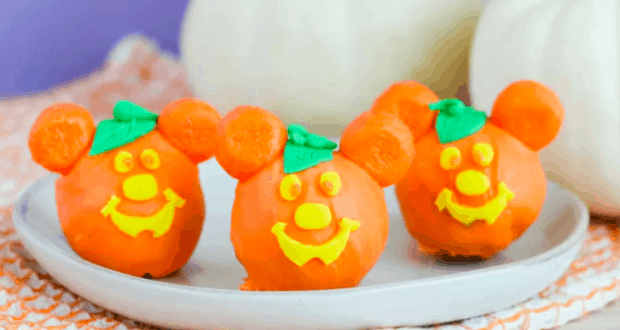 Three orange Mickey-shaped pumpkin truffles with cheerful yellow faces and green leaf accents are displayed on a white plate, making them the perfect festive treat for Halloween celebrations at Disney Parks.
