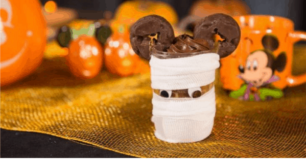 A mummy-wrapped cup with googly eyes and two chocolate donuts sits on a shimmering gold cloth, creating a whimsical Halloween treat display perfect for pairing with specialty cake jars or Mickey Mummy Cake Jars found at Disney parks during seasonal celebrations.