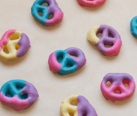 A tray of pastel-dipped pretzels in blue, pink, yellow, and purple candy coating is displayed at a themed food kiosk inspired by The Nightmare Before Christmas, offering whimsical Halloween treats reminiscent of snacks found at Disney Parks during the spooky season.