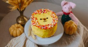Winnie the Pooh lion-inspired cake decorated with festive sprinkles, displayed alongside autumn pumpkins, bundles of wheat, and a cuddly pink plush at a whimsical character-themed event.