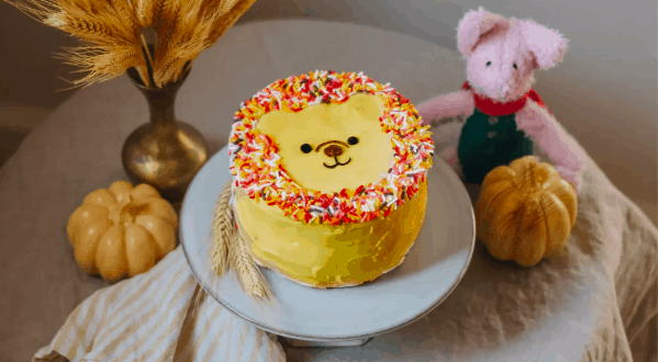 Winnie the Pooh lion-inspired cake decorated with festive sprinkles, displayed alongside autumn pumpkins, bundles of wheat, and a cuddly pink plush at a whimsical character-themed event.