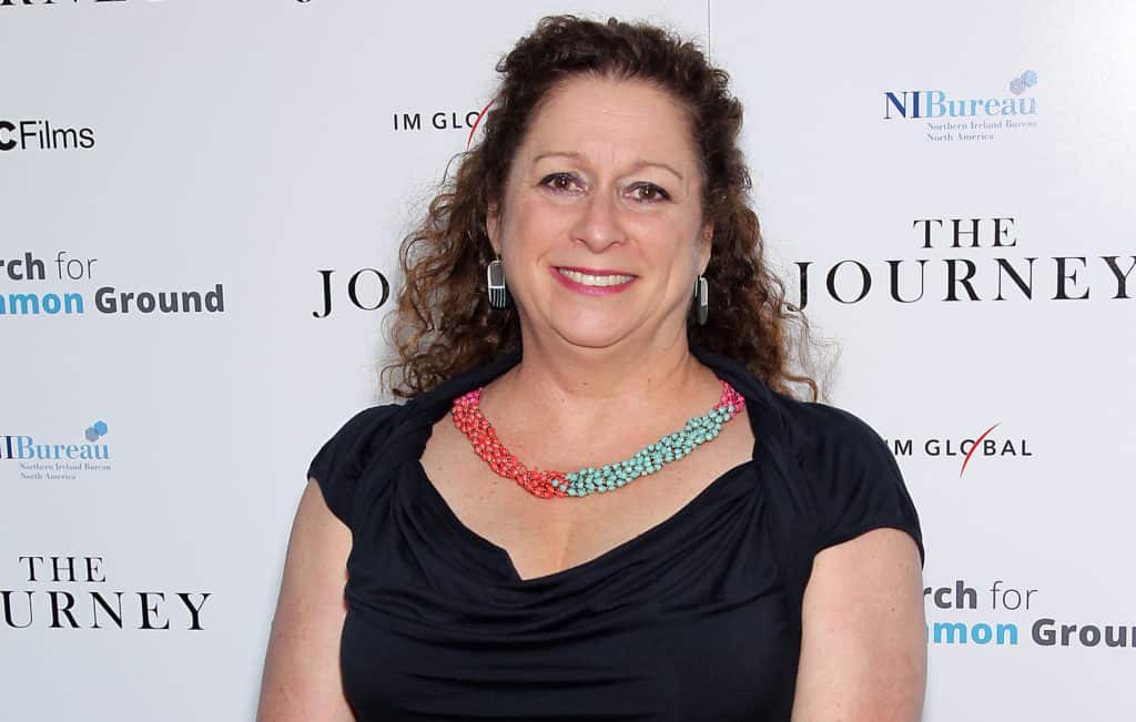 Abigail Disney, with curly hair, dressed in a black gown and vibrant necklace, beams in front of an elegant Disney event backdrop, radiating the signature magic and excitement of a Walt Disney Company celebration.