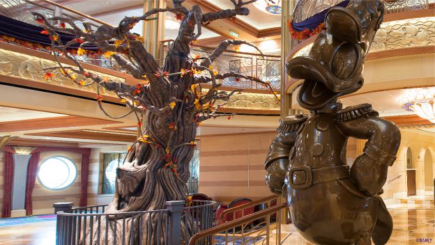 Donald Duck statue alongside a playful autumn Pumpkin Tree inspired by Disney Cruise Line, showcased within the lavish, storybook-style interior of a Disney park attraction.