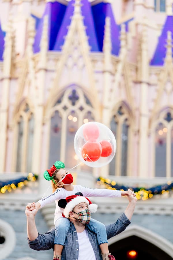 A young guest sporting Mickey ears and a mask beams atop an adult’s shoulders, clutching a festive balloon in front of Cinderella Castle—picture-perfect magic ready to be captured by Disney PhotoPass at Magic Kingdom.