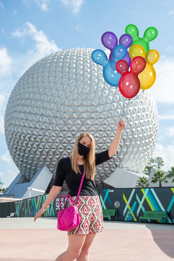 Disney fan posing in front of Epcot’s iconic Spaceship Earth, holding a bundle of colorful Mickey-shaped balloons, masked up and making memories with Disney PhotoPass.