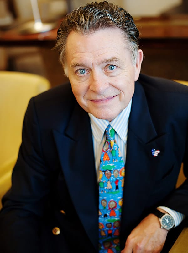 A cheerful former Disney executive in a sharp suit and vibrant tie, blue eyes sparkling, sits comfortably on a bright yellow chair, looking directly at the camera—poised to share behind-the-scenes stories and personal experiences from his adventures after leaving the Disney parks magic.