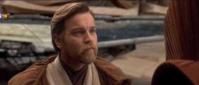 Obi-Wan Kenobi, with his signature beard and brown Jedi robe, stands outdoors at the park, gazing intently at a guest as he brings the legendary Star Wars character to life.
