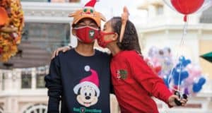 Guests in festive Disney holiday outfits enjoy a magical moment at Disney Parks, as one child gives their friend a sweet cheek kiss while holding colorful Disney balloons—perfect for capturing the enchantment and joy of the season.