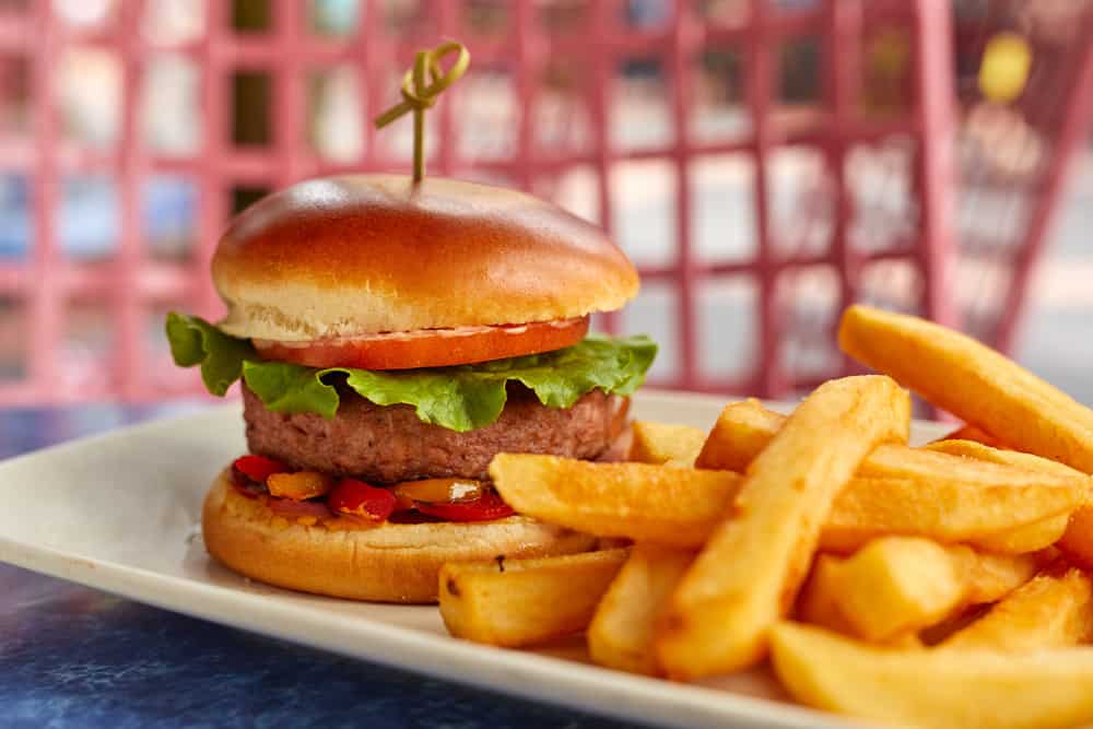 Dig into a mouthwatering new burger loaded with fresh lettuce and tomato on a soft bun, paired with golden thick-cut fries—all served on a white plate as part of the latest flavor-packed lineup at Disney’s Hollywood Studios.