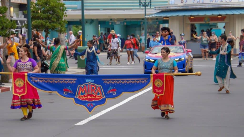 Mira Royal Detective" leads a vibrant parade through Disney's Hollywood Studios, with colorful performers and a festive car, as energetic Disney Junior entertainers engage the crowd.