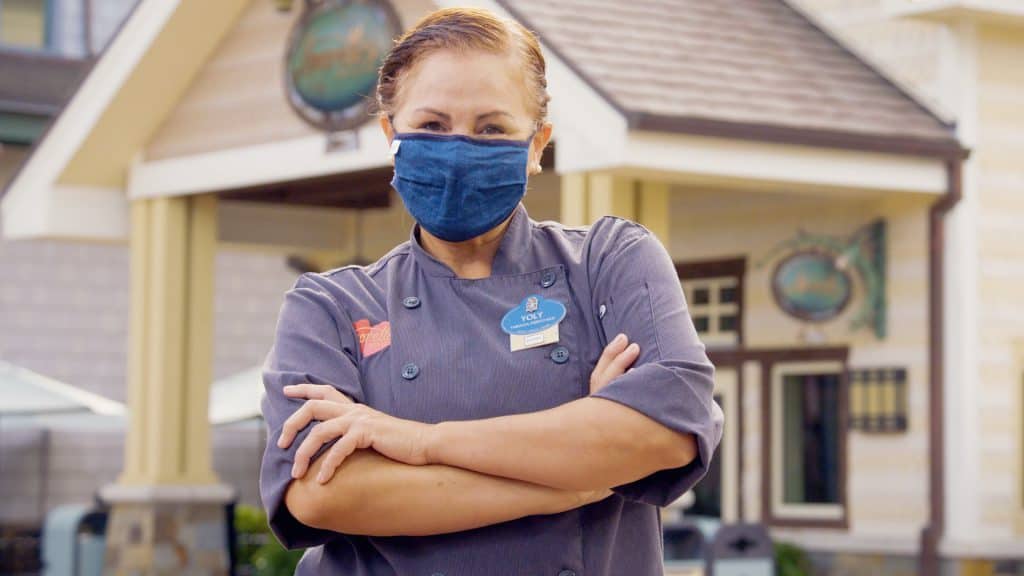 A Disney chef in a blue mask and uniform stands confidently outside a building at Walt Disney World, arms crossed and smiling for the camera, proudly showcasing Hispanic heritage and bringing Cultura & Sabor to Disney fans.