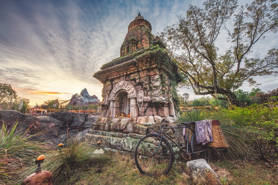 An ancient stone shrine draped in lush vines stands beside a bicycle, glowing in the golden sunset with trees and mountains behind—a scene straight from an immersive Memory Maker adventure, capturing the wonder and timeless magic you’d find exploring a storybook corner of your favorite theme park.