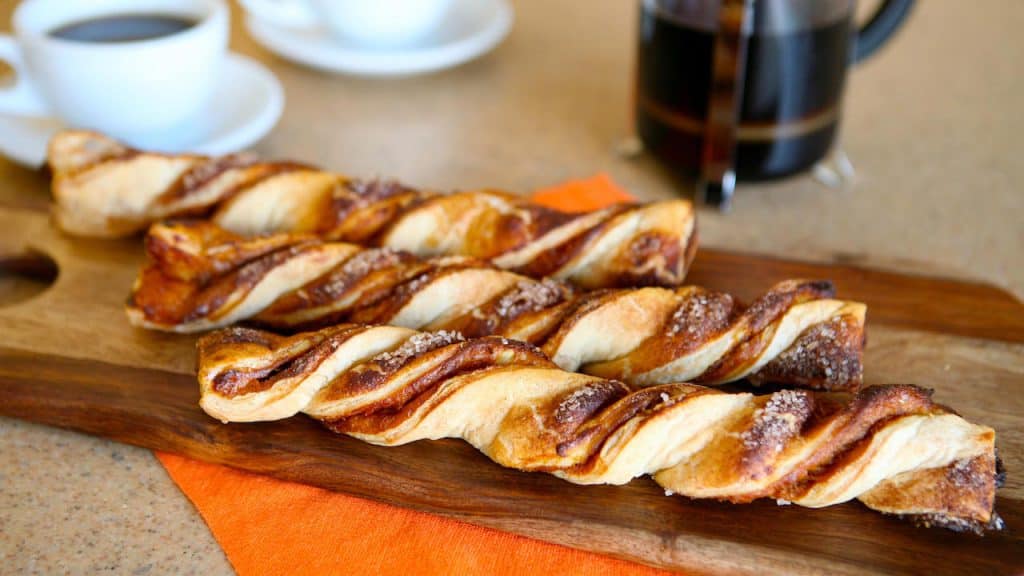 Pumpkin-twist pastries displayed on a rustic wooden board, surrounded by themed coffee cups and an orange napkin, inviting guests to enjoy a festive snack break between rides at the park—ideal for fans seeking seasonal treats and cozy fall flavors during the National Pumpkin Day celebration.
