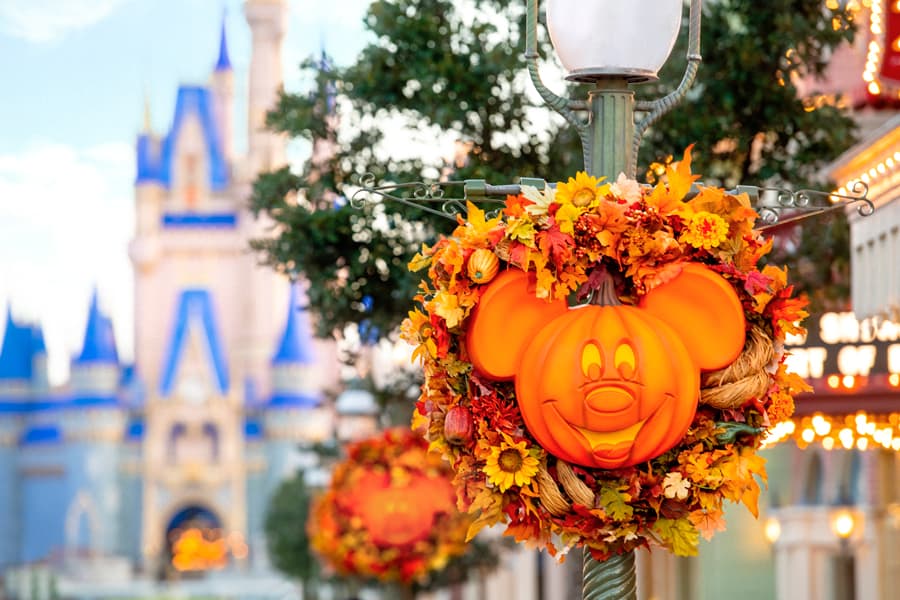 A festive orange Mickey pumpkin wreath adorned with autumn leaves decorates a streetlamp along Main Street, U.S.A., with Cinderella Castle softly blurred in the background—an enchanting fall scene perfect for capturing with Disney PhotoPass to create magical memories at Walt Disney World Resort.