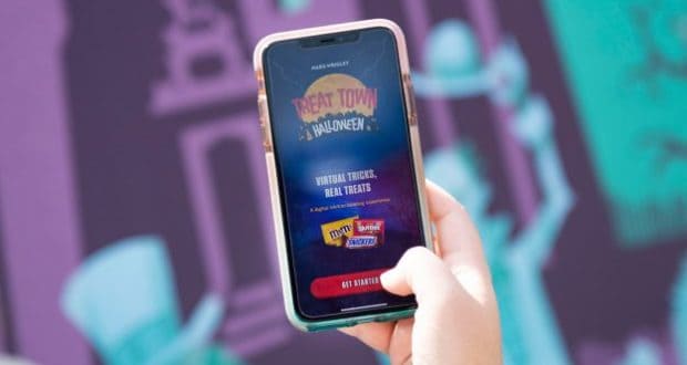 A guest’s hand holds a smartphone displaying the “TREAT TOWN Halloween” app, featuring beloved candy brands, a tribute to Disney’s The Haunted Mansion, and an inviting "Get Started" button—capturing the spirit of Disney Parks’ Halloween celebrations.