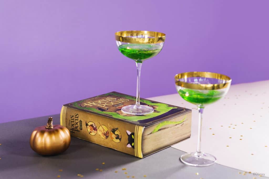 Two gold-rimmed glasses filled with vibrant green drinks inspired by magical Disney potions sit on a table alongside a Hocus Pocus spellbook and a shimmering gold pumpkin, channeling the whimsical Halloween atmosphere of the Sanderson Sisters from Disney’s classic film.