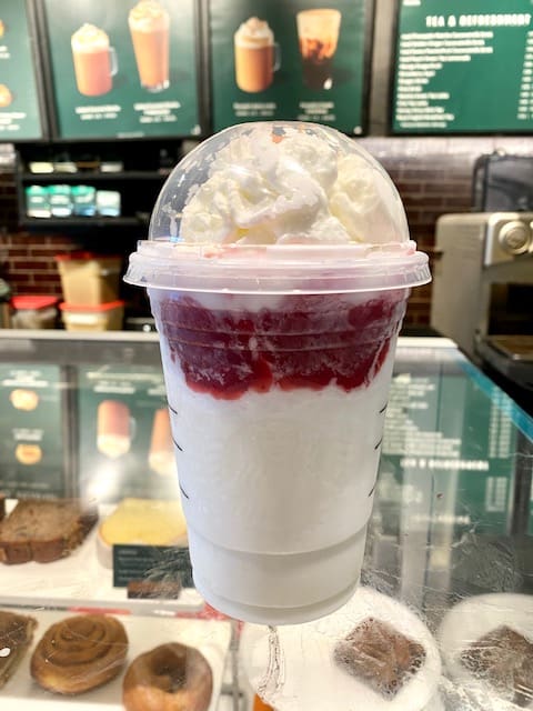 A vibrant strawberry and cream blended drink topped with fluffy whipped cream, inspired by the Starbucks Secret Menu, rests on a café counter inside Universal Studios, with the themed menu in the background.