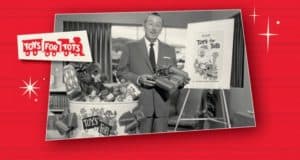 A cast member in a sharp suit stands next to a Toys for Tots collection bin and festive poster on a bright red backdrop at Disneyland Resort, holding gifts and encouraging guests to join in the spirit of giving this holiday season.