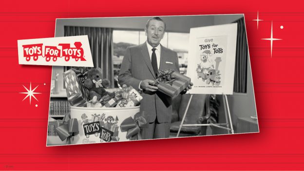 A cast member in a sharp suit stands next to a Toys for Tots collection bin and festive poster on a bright red backdrop at Disneyland Resort, holding gifts and encouraging guests to join in the spirit of giving this holiday season.