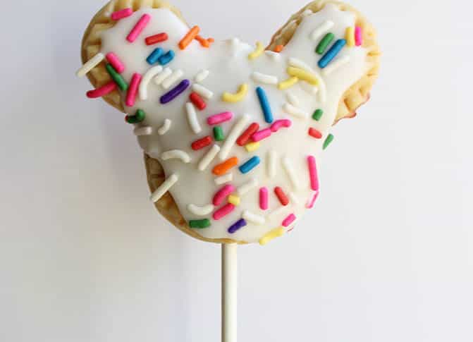 A whimsical Mickey Pie Pop shaped like Mickey Mouse, coated in white icing and topped with vibrant sprinkles, served on a stick—reminiscent of the magical snacks found at Disney parks and perfect for adding a touch of Disney magic to your next celebration.