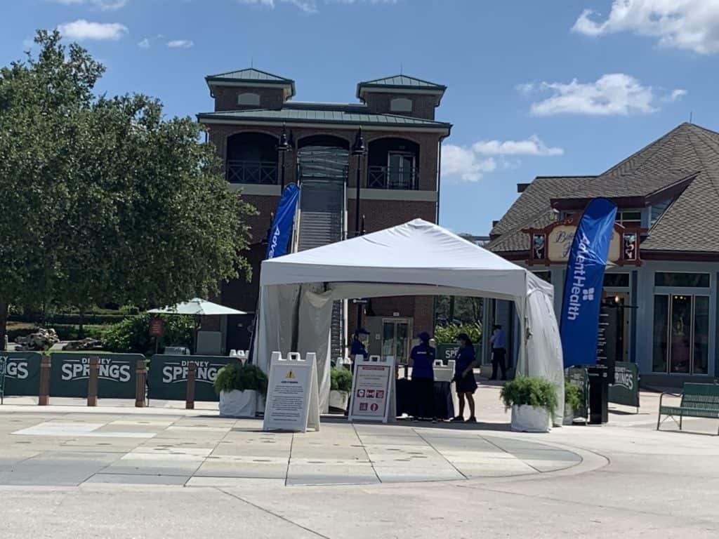 AdventHealth Orlando’s white vaccine tent set up amid cheerful blue Disney flags and a prominent health sign, inviting guests at Disney World’s sunny plaza near the resort buildings to easily access COVID-19 vaccines—an initiative supporting the well-being of visitors as part of Disney’s Health Partner commitment.
