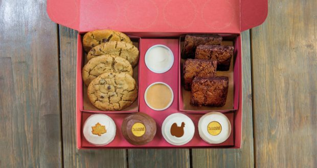 An open red box filled with chocolate chip cookies, brownies, and cupcakes topped with colorful sprinkles alongside sweet dips, set out on a wooden table—this Gratitude Bake Box is a magical assortment inspired by the irresistible desserts found at Disney Springs.
