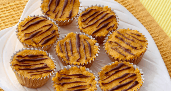 Eight allergy-friendly pumpkin muffins topped with chocolate drizzle, inspired by seasonal theme park treats, are artfully displayed on a white plate over cheerful yellow fabric—ready to delight fans of festive park snacks.