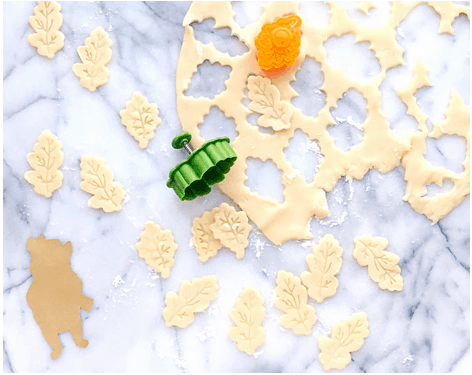 Leaf-shaped cookie dough cutouts and two cookie cutters sit on a marble countertop next to rolled-out dough, ready for baking treats inspired by your favorite theme park’s fall festivities—perfect for recreating sweet seasonal magic at home.