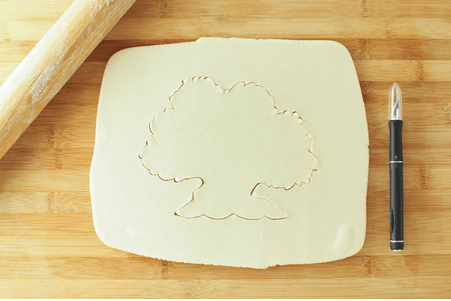 On a wooden surface, cookie dough is rolled out with a tree-shaped cutout, channeling the magic of Disney’s Animal Kingdom. A pen and rolling pin rest nearby, setting the stage for Disney fans to create their own Tree of Life Pie inspired by one of the park’s iconic landmarks.