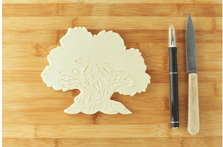 A Tree of Life pie dough cutout rests on a rustic wooden board, flanked by a peeler and serrated knife—channeling the creative spirit of Disney’s Animal Kingdom and inviting fans to craft their own culinary tribute to this iconic park centerpiece.