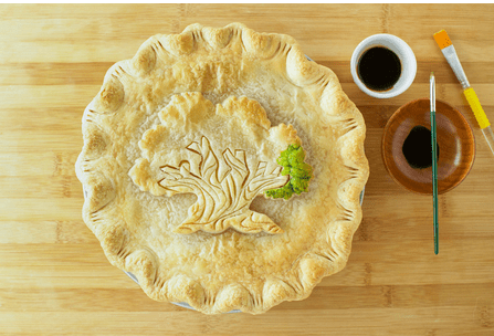 A golden pie featuring a crust design inspired by Disney’s Animal Kingdom’s Tree of Life, complete with a paintbrush, colorful sauce bowls, and fresh parsley garnish on a rustic wooden table—perfect for Disney fans and culinary adventurers.