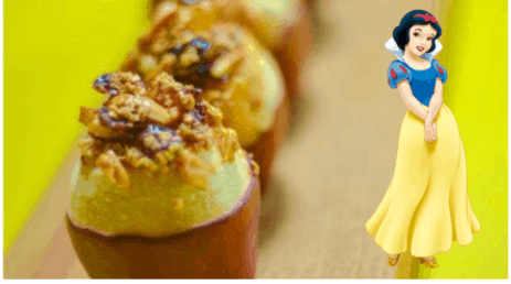 Baked apples with crumble topping on a cheerful yellow surface, inspired by the Snow White storybook recipe, with Snow White herself greeting guests beside the treats in a magical theme park setting.