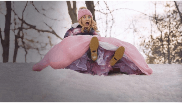 A wide-eyed girl in a pink dress and festive winter hat zips down a sparkling snowy hill beneath towering trees, evoking the wonder of December as if she's starring in a brand-new Disney+ adventure come to life at Disneyland.