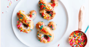 Three Mickey Mouse-shaped donuts covered in colorful sprinkles sit on a plate beside a cup of extra sprinkles, capturing the playful spirit of Disney parks—an irresistible treat for fans who crave magical snacks like Mickey-Shaped Pretzels at Walt Disney World or Disneyland.