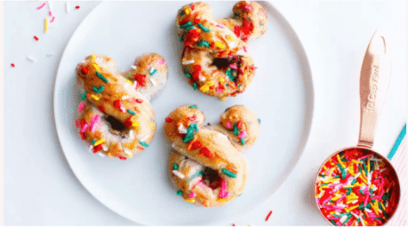 Three Mickey Mouse-shaped donuts covered in colorful sprinkles sit on a plate beside a cup of extra sprinkles, capturing the playful spirit of Disney parks—an irresistible treat for fans who crave magical snacks like Mickey-Shaped Pretzels at Walt Disney World or Disneyland.