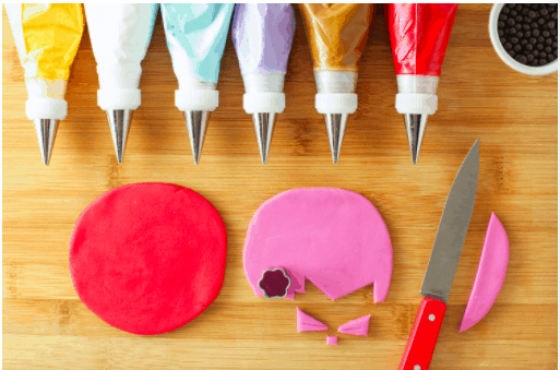 A colorful spread of six icing bags, a knife, black candy, and whimsical pink fondant shapes sits on a wooden board, all ready for crafting magical Mickey and Friends Icebox Cookies just like you’d find at Disney Parks.