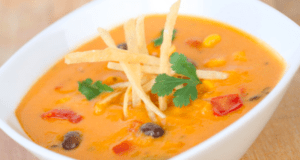 A vibrant bowl of Cheesy Enchilada Soup, swirling with bold orange cheese, black beans, red peppers, crispy tortilla strips, and a sprinkle of cilantro—served up like fan-favorite park fare ready to fuel your adventure between rides.