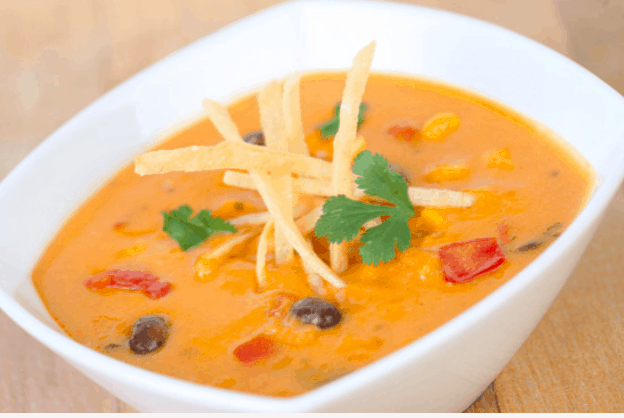 A vibrant bowl of Cheesy Enchilada Soup, swirling with bold orange cheese, black beans, red peppers, crispy tortilla strips, and a sprinkle of cilantro—served up like fan-favorite park fare ready to fuel your adventure between rides.